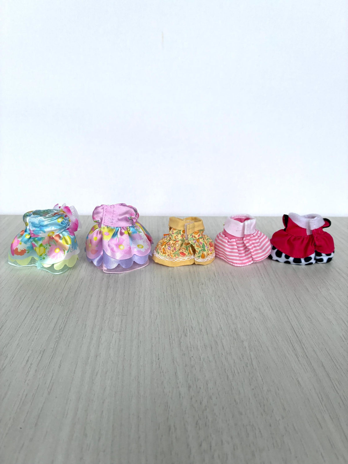 Calico Critters and Sylvanian Families child-size dresses shown together in a you-pick group photo: two Flowering Princess floral ruffle gowns, a yellow floral dress with an orange bow, a pink and white striped dress, and a black-and-white Dalmatian polka dot dress.