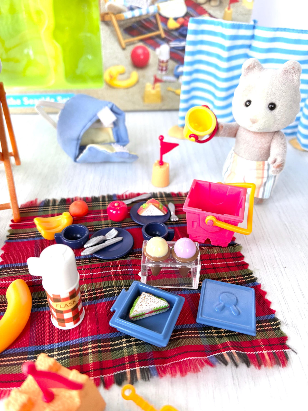 Sylvanian Families Calico Critters Day at the Seaside Set (Flair 4870): close-up of the picnic rug with flask, ice cream box, sandwich box with lid, plates, cutlery and red bucket