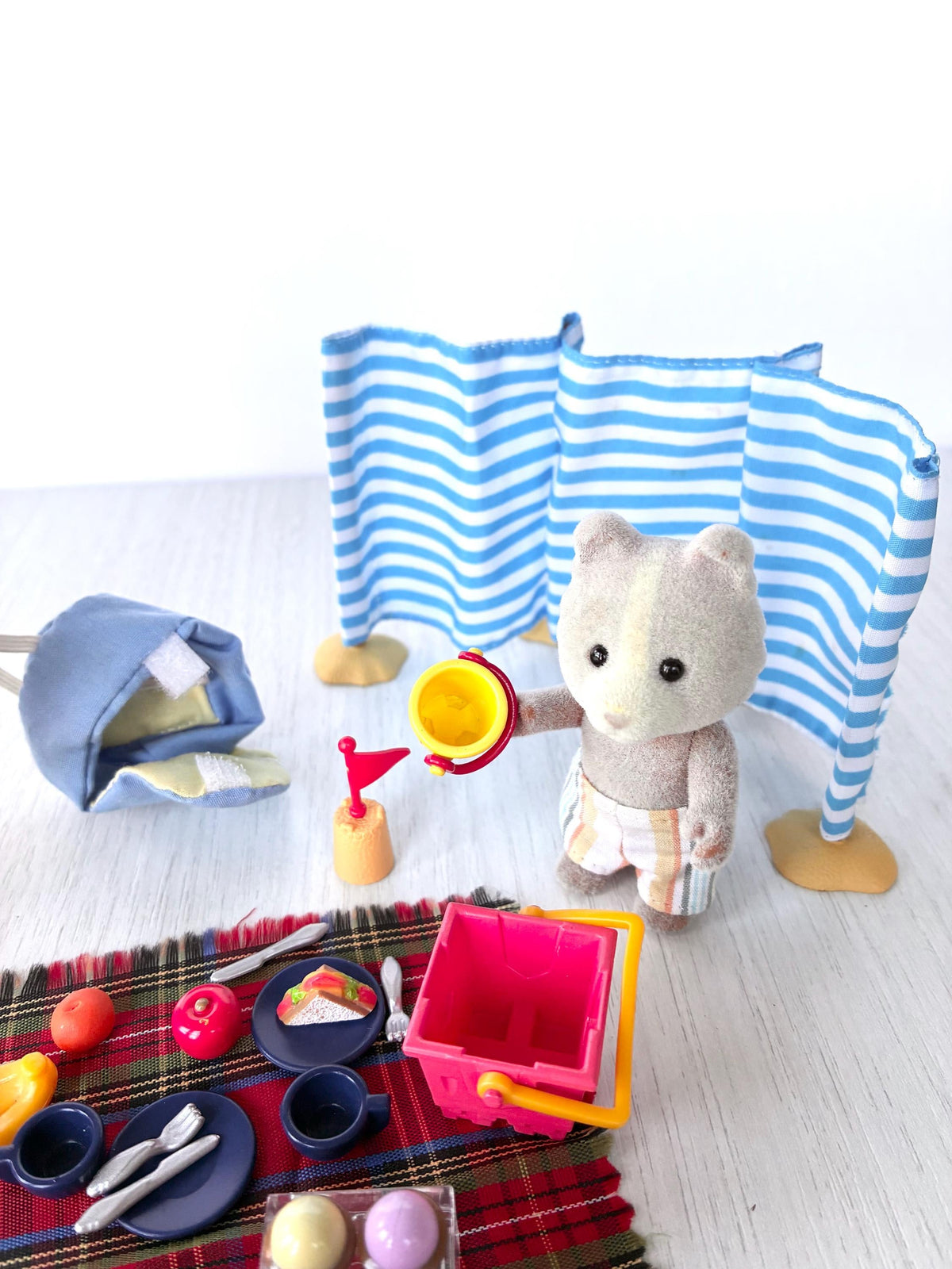 Sylvanian Families Calico Critters Day at the Seaside Set (Flair 4870): Vespa Farthing holding the yellow bucket by the rug with plates, cutlery, fruit and red bucket