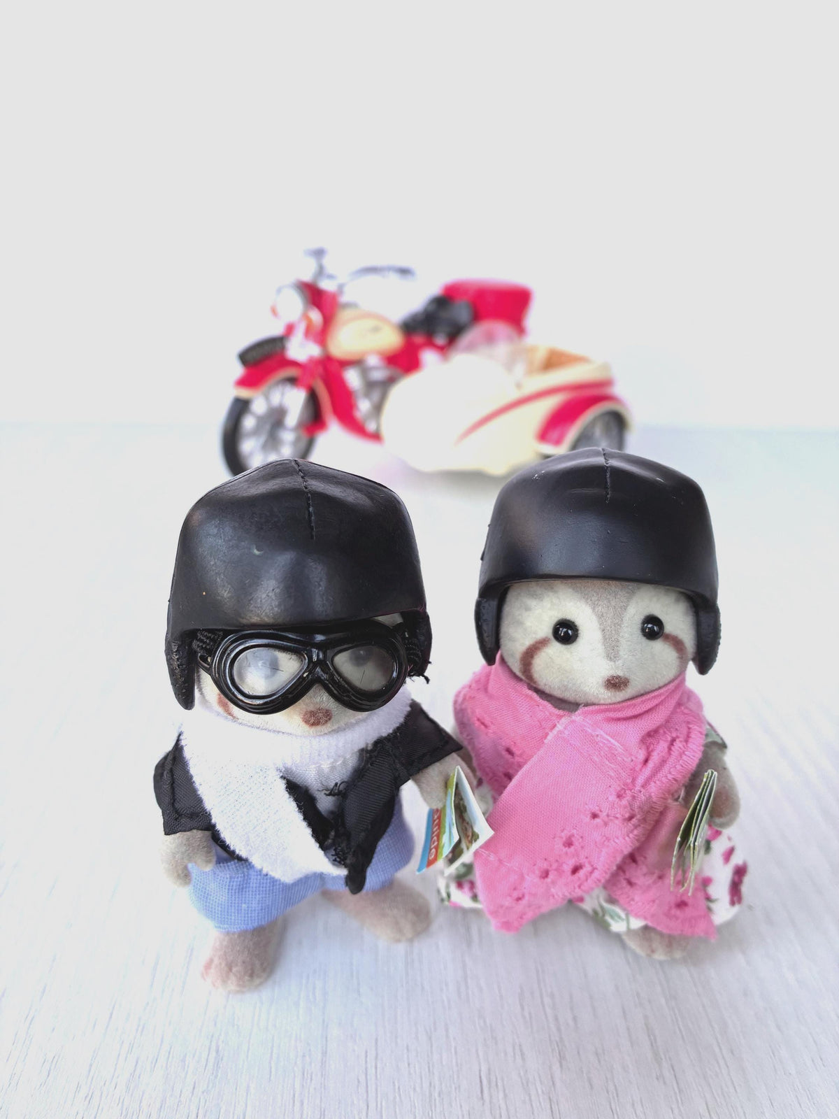 Calico Critters Sylvanian Families Mulberry Raccoon Grandparents wearing black riding helmets, George in goggles and Mildred in pink shawl, motorcycle behind them