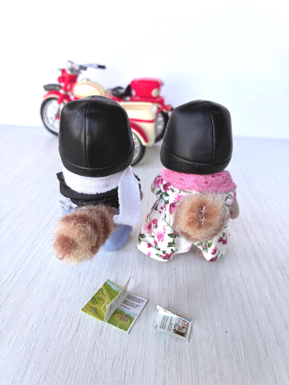 Calico Critters Sylvanian Families Mulberry Raccoon Grandparents from behind showing striped raccoon tails and helmets, open map and guide on the table