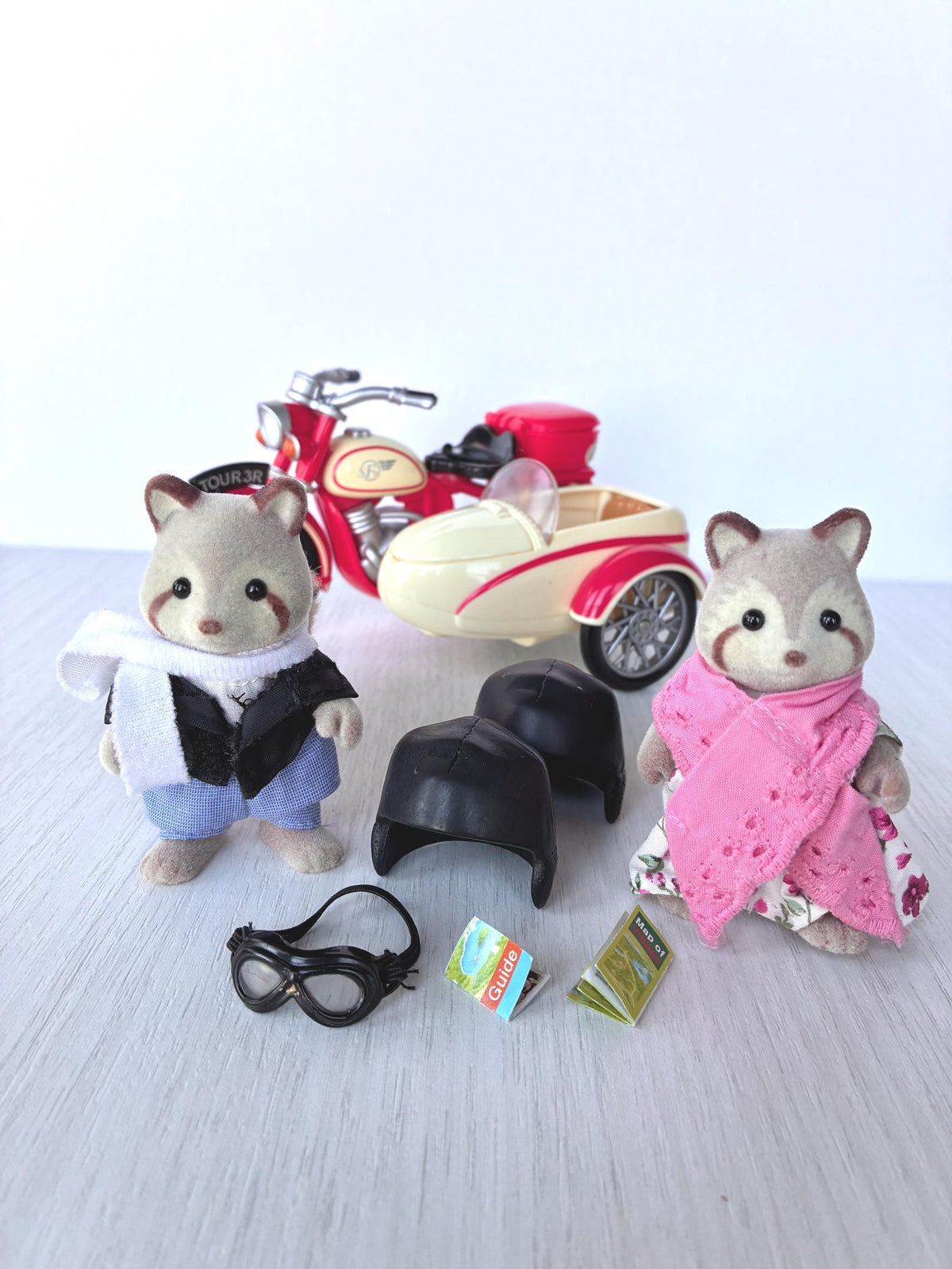 Calico Critters Sylvanian Families Mulberry Raccoon Grandparents standing beside the red Motorcycle and Sidecar with their two black helmets, goggles, village guide and map